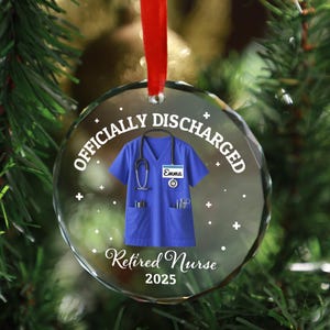 Könnte beinhalten: Ein klarer, runder Weihnachtsschmuck mit rotem Band. Der Schmuck zeigt ein blaues Krankenschwester-Uniform-Design mit Stethoskop und den Texten "Officially Discharged" und "Retired Nurse 2025". Der Name "Emma" steht auf der Uniform.