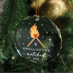 Könnte beinhalten: Ein klarer, runder Weihnachtsbaumschmuck aus Glas mit einer goldenen Schnur. Der Schmuck zeigt eine Grafik von zwei Streichhölzern mit einer Flamme und einem Herzen, die Namen "SOPHIA & MATYHEW", das Wort "matched" und das Jahr "2025".