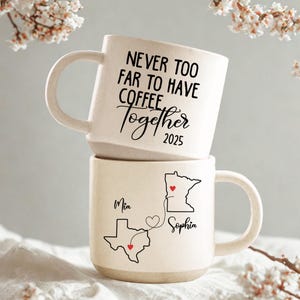 Könnte beinhalten: Zwei gestapelte cremefarbene Keramik-Kaffeetassen. Die obere Tasse trägt den Text "NEVER TOO FAR TO HAVE COFFEE Together 2025". Die untere Tasse zeigt eine Karte von Texas und Minnesota mit den Namen "Mia" und "Sophia".