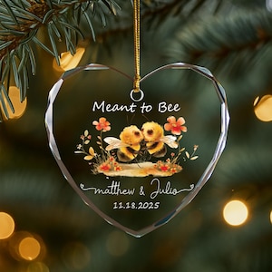 May include: Heart-shaped glass ornament with a gold string, featuring two cartoon bees kissing. The ornament has the text "Meant to Bee", names "matthew & Julia", and the date "11.18.2025". Decorative flowers surround the bees.