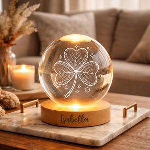 Könnte beinhalten: Eine klare Glaskugel mit einem weißen, geätzten Kleeblatt-Design, von innen beleuchtet, steht auf einem Holzsockel. Der Sockel ruht auf einem Marmortablett mit goldenen Griffen. Der Name "Isabella" ist auf dem Holzsockel eingraviert. Eine brennende Kerze und dekorative Gegenstände befinden sich im Hintergrund.