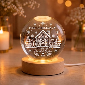May include: A clear glass sphere night light on a round wooden base. The sphere features white etched designs of a gingerbread house, gingerbread figures, trees, snowflakes, and a star. Text reads: FIRST CHRISTMAS AT 12 Holly Lane Daniel & Sophia 2026.