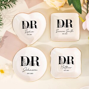 Könnte beinhalten: Vier weiße Keramik-Schmuckschalen mit goldenen Rändern. Jede Schale hat eine andere Form: Herz, Quadrat, rund und quadratisch mit Zacken. Jede Schale zeigt die Initialen "DR" mit floralem Design, einem Namen und "EST. 2025".