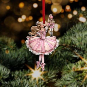 Puede incluir: Adorno navideño de bailarina rosa y blanca con un cascanueces a juego. La bailarina lleva un tutú rosa y zapatillas de ballet. El adorno cuelga de una cinta roja sobre un fondo borroso de ramas de árbol de Navidad.