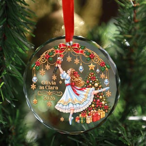 May include: A round, faceted glass Christmas ornament hanging from a red ribbon. The design features a girl in a white dress holding a nutcracker, a decorated Christmas tree with gifts, gold snowflakes, and the text Olivia as Clara 2026.