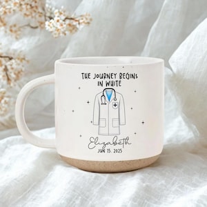 Könnte beinhalten: Weißer Keramikbecher mit gesprenkeltem Boden und gebogenem Henkel. Der Becher zeigt den Text "THE JOURNEY BEGINS IN WHITE" und eine Grafik eines Arztmantels und eines Stethoskops. Der Name "Elizabeth" und das Datum "JUN 15. 2025" sind ebenfalls aufgedruckt.
