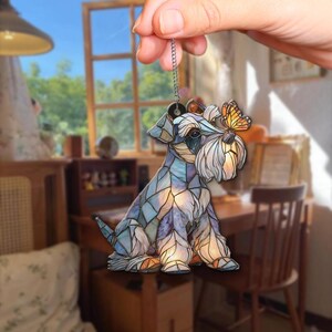 May include: A decorative Schnauzer dog ornament with a stained glass effect. The dog is primarily blue and purple, with a white beard and a small orange butterfly perched on its nose. It hangs from a silver chain.