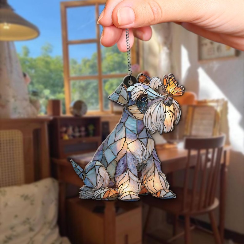 May include: A decorative Schnauzer dog ornament with a stained glass effect. The dog is primarily blue and purple, with a white beard and a small orange butterfly perched on its nose. It hangs from a silver chain.