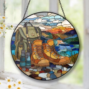 Könnte beinhalten: Runde Glasmalerei mit einer Wanderszene. Das Kunstwerk zeigt einen grünen Rucksack, braune Wanderstiefel und ein Fernglas. Der Hintergrund zeigt eine farbenfrohe Landschaft mit Bergen, Wasser und Sonnenuntergang. Die Tafel ist schwarz gerahmt und hängt an einer Kette.
