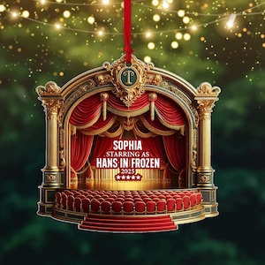 Op de afbeelding: Een kerstversiering met theaterthema en een rood lint. De versiering toont een podium met rode gordijnen, gouden accenten en de tekst "SOPHIA STARRING AS HANS IN FROZEN 2025".