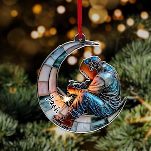 May include: A Christmas ornament featuring a welder in a blue and orange outfit, seated on a crescent moon. The moon is made of stained glass with various colors. The name "Jack" is written on the ornament.