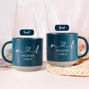Könnte beinhalten: Zwei tealfarbene Keramikbecher mit weißem Text und grauem Boden. Der Text auf den Bechern lautet "mel" mit einem Herzsymbol, "ENGAGED" und dem Datum "17.08.25". Die Becher haben einen gebogenen Griff.