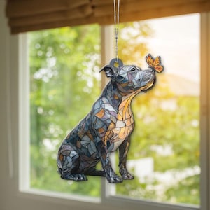 Op de afbeelding: Een decoratief hangornament met een zittende hond in glas-in-loodstijl. De hond is voornamelijk blauw, grijs en oranje, met een vlinder op zijn neus. Het hangt aan een zilveren ketting.
