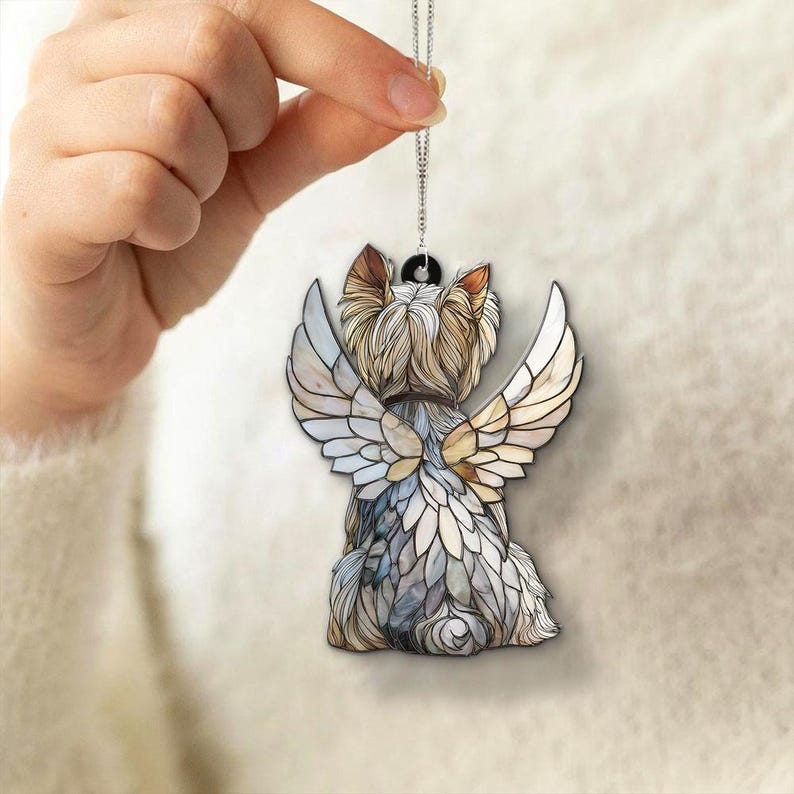 May include: A pendant depicting a Yorkshire Terrier with angel wings. The dog is seen from the back, with detailed fur and feather-like wings in shades of white, beige, and grey. The pendant hangs from a silver chain.