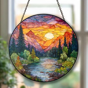 Könnte beinhalten: Runde Glasmalerei mit einem Sonnenuntergang über einer Bergkette und einem Fluss. Das Kunstwerk zeigt leuchtende Farben wie Orange, Gelb, Blau und Grün, mit einem schwarzen Metallrahmen und einer Kette zum Aufhängen. Die Szene umfasst Bäume, Wasser und eine helle Sonne.