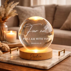 Könnte beinhalten: Eine leuchtende Glaskugel mit dem Text "Fear not, FOR I AM WITH YOU, Isaiah 41:10" und dem Namen "Sophia". Die Kugel steht auf einem Holzsockel auf einem Marmortablett mit goldenen Griffen. Eine brennende Kerze und eine dekorative Vase sind im Hintergrund.