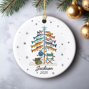 May include: White ceramic Christmas ornament with a fishing theme. The ornament features a Christmas tree made of colorful fish, a fishing rod, a tackle box, and a small green chair. The name "Jackson" and the year "2025" are printed on the ornament.
