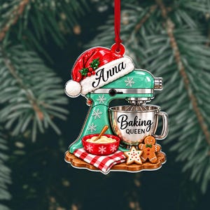 May include: A flat Christmas ornament shaped like a mint green stand mixer wearing a red Santa hat with the name Anna. It features a silver mixing bowl labeled Baking Queen, a red bowl with dough, a gingerbread man, and a star cookie.