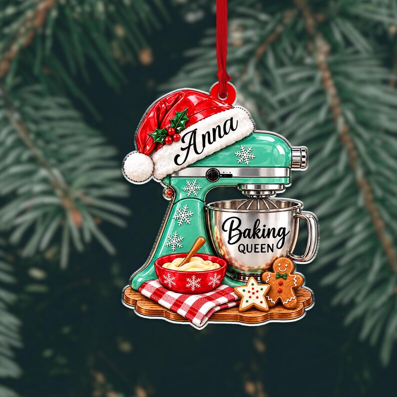 May include: A flat Christmas ornament shaped like a mint green stand mixer wearing a red Santa hat with the name Anna. It features a silver mixing bowl labeled Baking Queen, a red bowl with dough, a gingerbread man, and a star cookie.
