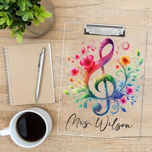 May include: A clear clipboard with a silver clip, featuring a colourful treble clef design with flowers and the name "Mrs. Wilson" in black script. The design includes pink, red, orange, and blue hues.
