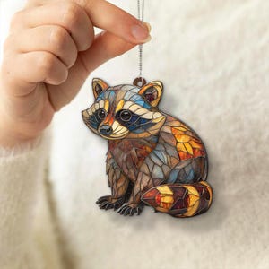 May include: A colorful raccoon ornament with a stained-glass effect. The raccoon is depicted in shades of brown, orange, yellow, blue, and gray. It is hanging from a silver chain. The ornament is held by a hand.