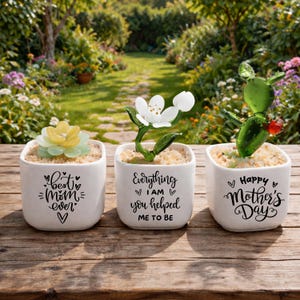 Könnte beinhalten: Drei weiße quadratische Keramik-Pflanzgefäße mit dekorativen Glasblumen. Die Pflanzgefäße tragen die Aufschriften "Best Mom Ever", "Everything I am you helped me to be" und "Happy Mother's Day". Die Pflanzgefäße stehen auf einer Holzoberfläche.