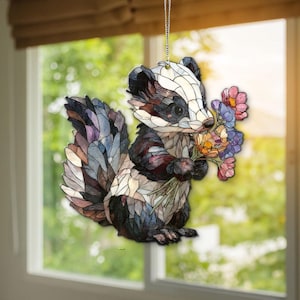 May include: A stained-glass effect ornament of a badger holding a bunch of flowers. The badger has a black, white, and brown coat, and the flowers are pink, purple, and orange. The ornament hangs from a silver chain, suitable for a window.