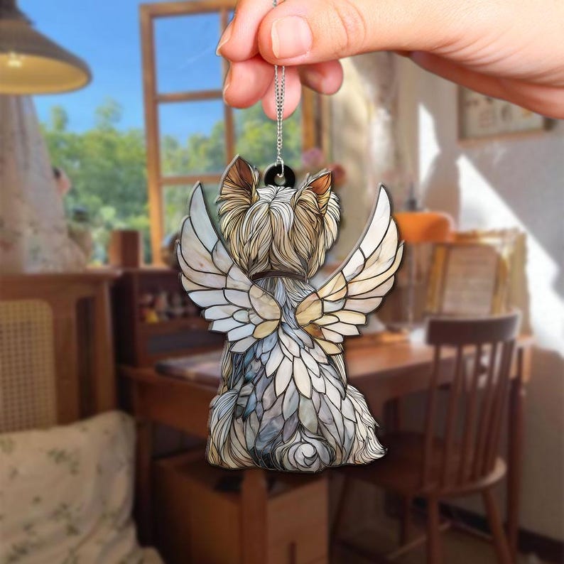 May include: A pendant depicting a Yorkshire Terrier dog with angel wings. The dog has tan and white fur, and the wings are white and beige. The pendant is suspended from a silver chain. The background is a blurred interior scene.