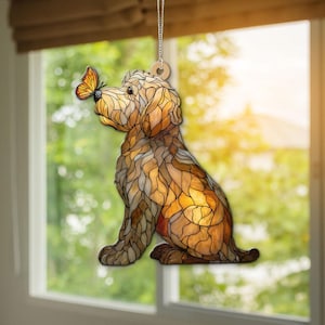 Op de afbeelding: Een decoratief hondenornament met een glas-in-lood effect. De hond is bruin en bruingeel, met een vlinder op zijn neus. Het ornament hangt aan een zilveren ketting en een houten schijf.