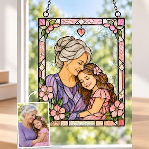 Könnte beinhalten: Ein rechteckiger Buntglas-Sonnenfänger mit einer Illustration einer älteren Person mit grauen Haaren in einem violetten Oberteil, die ein kleines Kind in einem rosa Kleid umarmt. Der Rand ist mit rosa Blumen und einem kleinen Herz verziert.