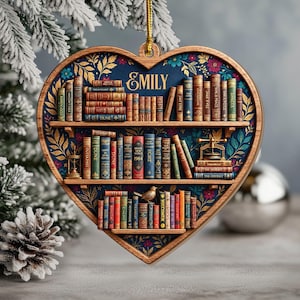 Könnte beinhalten: Ein herzförmiger Holzschmuck mit dunkelblauem Hintergrund, Blumenmustern und drei Regalen mit Miniaturbüchern. Der Name Emily ist oben aufgedruckt. Die Regale enthalten verschiedene Buchrücken mit Titeln wie Dune, 1984 und The Hobbit.