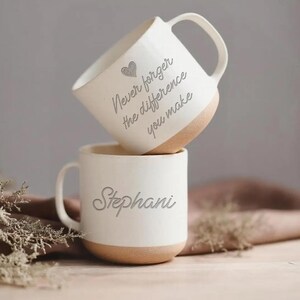 May include: Two white ceramic mugs with a tan base are stacked. The top mug has the text "Never forget the difference you make" and a heart. The bottom mug has the name "Stephani".