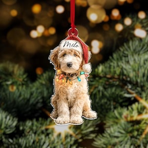 May include: A Christmas ornament featuring a light brown dog wearing a Santa hat with the name "Milo" and a string of colorful lights. The ornament is hanging from a red ribbon against a blurred background of Christmas tree branches.