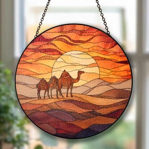 Könnte beinhalten: Runde Glasmalerei mit einer Wüstenszene, die drei Kamele vor einem Sonnenuntergang zeigt. Das Kunstwerk verwendet warme Farbtöne von Orange, Rot und Braun, mit einer schwarzen Kette zum Aufhängen. Das Design erinnert an eine Wüstenlandschaft.