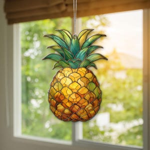 Könnte beinhalten: Hängende Ananas-Dekoration mit Glasmalerei-Effekt. Der Ananas-Körper ist gelb und orange, die Blätter grün. Sie hängt an einer silbernen Kette und erzeugt ein tropisches Ambiente.
