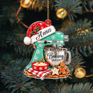 May include: A flat Christmas ornament shaped like a mint green stand mixer wearing a Santa hat with the name Anna. The mixer bowl features the text Baking Queen. A red bowl with dough, a red and white checkered cloth, and gingerbread cookies sit below.