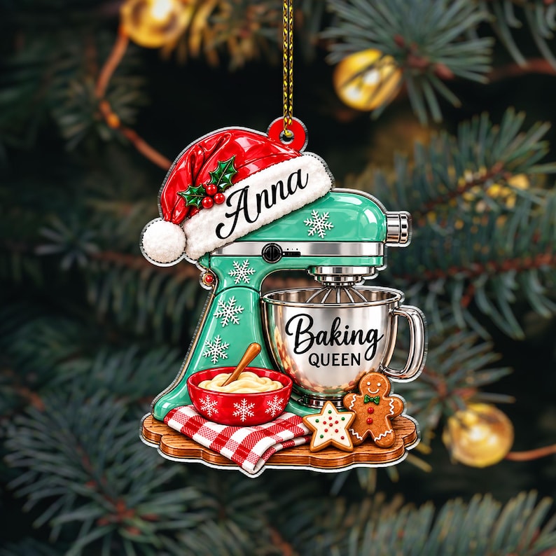 May include: A flat Christmas ornament shaped like a mint green stand mixer wearing a Santa hat with the name Anna. The mixer bowl features the text Baking Queen. A red bowl with dough, a red and white checkered cloth, and gingerbread cookies sit below.
