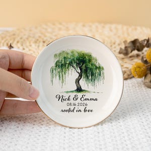 Könnte beinhalten: Eine weiße Keramik-Ringschale mit Goldrand. Die Schale zeigt ein Aquarellgemälde einer Trauerweide. Der Text "Nick & Emma 08.16.2026 rooted in love" ist unter dem Baum aufgedruckt.