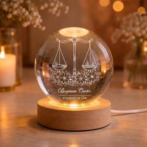 Könnte beinhalten: Eine klare Glaskugel mit einem weißen, gravierten Waage-der-Gerechtigkeit-Design, floralen Akzenten und dem Text "Benjamin Carter, Attorney at Law". Die Kugel steht auf einem Holzsockel, der warm leuchtet.
