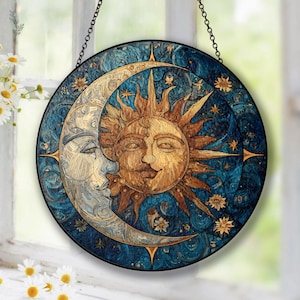 May include: Circular stained glass artwork depicting a sun and moon design. The sun has a smiling face, and the moon is a crescent with a face. The background is blue with stars. It is framed in black and suspended by a chain.