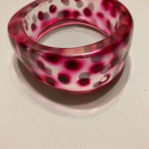 May include: A thick, translucent bangle bracelet made of clear resin. The bracelet features a repeating pattern of embedded dark magenta and soft grey circular spots throughout the material, creating a layered, multi-dimensional appearance.