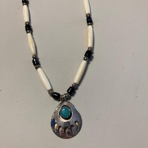 May include: A necklace featuring a silver-toned pendant shaped like a bear claw with a central turquoise stone. The cord is strung with alternating white cylindrical beads, small black spherical beads, and metallic spacers in a repeating pattern.