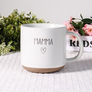 Puede incluir: Taza de cerámica blanca con la palabra "MAMMA" y un símbolo de corazón. La taza tiene una base marrón y un asa gris. La taza está sobre una superficie blanca con vegetación y flores rosas en el fondo.