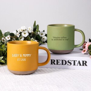 May include: Two ceramic mugs. One is mustard yellow with "DADDY & MOMMY EST.2026" in white text. The other is green with "Maybe coffee is addicted to me" in white text. Both have brown bases and large handles.