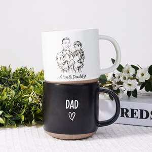 May include: Two ceramic mugs stacked on top of each other. The top mug is white with a black and white illustration of a man holding a child, with the text "Alan & Daddy". The bottom mug is black with the word "DAD" and a heart.