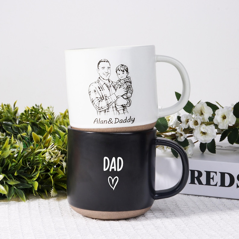 May include: Two ceramic mugs stacked on top of each other. The top mug is white with a black and white illustration of a man holding a child, with the text "Alan & Daddy". The bottom mug is black with the word "DAD" and a heart.