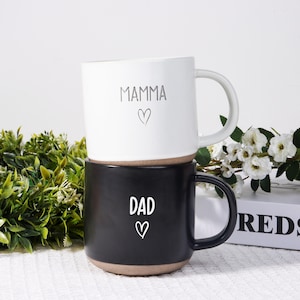 May include: Two ceramic mugs, one stacked on top of the other. The top mug is white with the word "MAMMA" and a heart symbol. The bottom mug is black with the word "DAD" and a heart symbol. Both mugs have a brown base and white handles.