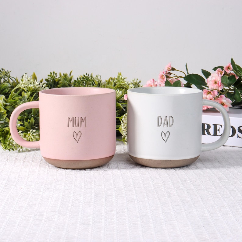 May include: Two ceramic mugs, one pink and one white, with brown bases. The pink mug says "MUM" with a heart, and the white mug says "DAD" with a heart. Both mugs have a handle and are on a white surface.