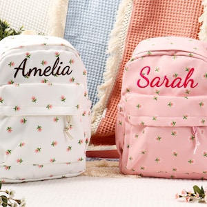 以下が含まれることがあります： 花柄のバックパック2つ。1つは白で「Amelia」が茶色で刺繍され、もう1つはピンクで「Sarah」がピンクで刺繍されています。どちらも小さなピンクの花のディテールとサイドポケットが付いています。