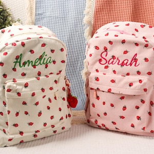 Può includere: Due zaini in velluto a coste, uno color crema e uno rosa, sono ricoperti da motivi a fragole rosse. Lo zaino color crema ha "Amelia" ricamato in verde, mentre quello rosa dice "Sarah". Ognuno ha una tasca frontale e un ciondolo a forma di fragola.